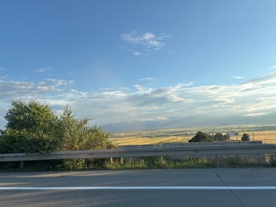 This media file depicts a scenic view from a roadside, likely a highway, looking out over a vast, agricultural valley. The sky is a brilliant blue, with scattered white clouds, suggesting it is daytime, possibly late afternoon given the soft, warm light illuminating the landscape. In the foreground, a silver guardrail separates the roadway from the elevated terrain, where lush green trees and vegetation are visible.
The main subject of the image is the expansive landscape beyond the guardrail. It appears to be a rural area with fields of golden crops, possibly wheat or hay, stretching towards a distant mountain range. The mountains are hazy and shrouded in a light mist, adding depth and a sense of tranquility to the scene. Scattered throughout the valley are various structures, including what seem to be farm buildings, industrial facilities, and utility poles, hinting at human activity within this natural setting. A single lamppost is visible in the middle ground.
The overall mood is peaceful and serene. The composition, with the clear sky, rolling fields, and distant mountains, evokes a sense of openness and natural beauty. The presence of the guardrail and the paved road in the foreground firmly places the viewer as an observer from a moving vehicle, perhaps enjoying a journey through the countryside. There is no visible text in the media file. The location context provided is Kvemo Shavshvebi, Georgia, which aligns with the rural, mountainous landscape.