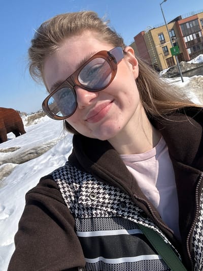 A young Caucasian female is depicted in a selfie, smiling slightly and wearing brown-framed sunglasses with reflective blue lenses. Her light brown hair is pulled back. She wears a brown jacket featuring black and white houndstooth patterns on the upper portion and horizontal black and white stripes below, over a light pink shirt. A dark green strap, likely from a bag, is visible on her right side. The scene is outdoors in Новое Шигалеево, Russia, under a clear blue sky, with patches of melting snow covering the ground. In the left background, a large, woven, brown bear sculpture is partially visible. To the right, a modern multi-story building with yellow and brown facades stands, alongside a street light pole with a small green sign attached.