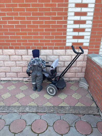 In Gamovo, Russia, a young child, viewed from behind, stands outdoors on a paved surface next to a building. The child wears a dark blue knit hat, a plaid long-sleeve top with blue, green, and white patterns, gray overalls, dark gloves, and dark boots. To the child's right is a dark-framed tricycle-stroller with three wheels, a light gray fabric canopy, and black handlebars; the right handlebar's foam grip is damaged. The ground is paved with two distinct sections: the foreground features gray and reddish circular pavers, while the area nearer the wall consists of red and light yellow/beige clover-shaped pavers. The wall behind the child and stroller is constructed with a lower course of light-colored, possibly tan, stone blocks, topped by red bricks. A vertical strip of white bricks is integrated into the red brick section on the right side of the frame. A small stack of gray paving stones is visible on the ground in the lower right corner, adjacent to the wall. The scene is illuminated by natural daylight.