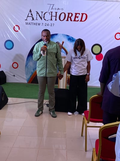A medium shot captures a man speaking into a microphone, standing alongside a woman wearing a t-shirt that says "ANCHORED." They are standing in front of a banner also bearing the word "ANCHORED," with the scripture reference Matthew 7:24-27 below. The banner design includes abstract circular shapes of various colors. The location is likely a church or event space in Miango, Nigeria, based on the setting. There are chairs and other people visible in the background.