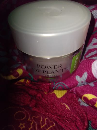 A close-up, overhead shot captures a jar of "POWER OF PLANTS OPUNTIA" cream, its lid a matte white and its container transparent, revealing the product inside. The jar is nestled within a vibrant fuchsia-colored fabric with a repeating floral pattern in white, yellow, and teal. To the right of the jar, a bright green sticker with the word "lirene" printed in white is visible. The fabric also features a fringe of deep red threads along the bottom edge of the frame. The lighting is dim, casting shadows and highlighting the textures of the fabric and the jar. The image is an indoor product shot with no indication of a specific location within Kitskany, Moldova.