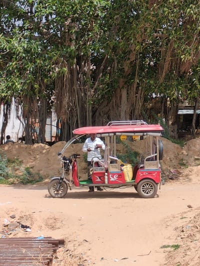 In Bandolai Ki Dhani, India, an outdoor scene unfolds beneath the sprawling branches of a large banyan tree. A man, dressed in a white shirt, stands beside a red and silver electric rickshaw. The rickshaw, which appears to be a passenger vehicle, is parked on a dirt road, with a sandy embankment behind it. The environment suggests a rural or semi-rural setting, with some vegetation visible and what appears to be construction materials or debris in the foreground. The overall lighting indicates daytime.