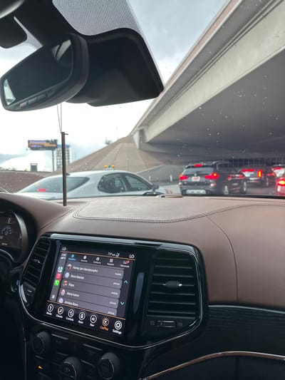 The image depicts the interior of a vehicle, likely in the City of Paradise, United States, looking forward through the windshield at a traffic-congested roadway. The vehicle's dashboard features brown leather stitching and black plastic trim. A central infotainment screen displays "4:06," "73° out," and a contact list including "Honey Jan HarutyunAn," "Rons Market," "Papa," "Alena Karen," and "Armine Rons," with options for calls. Below the screen, control buttons for "Climate," "Controls," "Apps," "Nav," "CarPlay," and "Settings" are visible. A rearview mirror assembly is visible at the top, with a dark, thin object hanging from it. The windshield has small particles, possibly dust or water droplets.
Outside the vehicle, a multi-lane road is populated with stationary or slow-moving vehicles under an overcast sky. A silver car is partially visible to the immediate left. Directly ahead, a black BMW SUV with illuminated red taillights is positioned. Further down the road, other vehicles are visible, positioned beneath a large, light-colored concrete overpass structure that spans overhead and to the right. In the far distance on the left, a digital billboard is discernible. No individuals are visible in the scene.