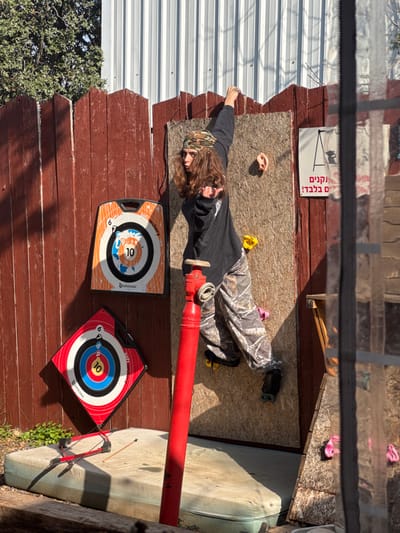 The image depicts an outdoor recreational area located within the Menashe Regional Council, Israel. A single individual, appearing young, with long wavy brown hair, wears a camouflage bandana, a black long-sleeved shirt, and camouflage pattern pants. This person is positioned on a vertical bouldering wall constructed from wooden particle board. Their right arm is extended upwards, gripping a climbing hold, while their left hand is forming a "rock on" gesture. Their right foot rests on a yellow climbing hold, and their left foot is on a pink climbing hold. The bouldering wall features multiple climbing holds, including yellow, pink, and a flesh-toned, ear-shaped hold.
To the left of the bouldering wall, a dark brown wooden fence with an uneven top edge extends across the background. Two square archery targets are affixed to this fence: an orange target with a blue, white, and orange bullseye, and a red target with a red, yellow, blue, black, and white bullseye. Both targets display "Soft Archery" and "Geologic" branding, along with numerical scoring zones. Below these targets, a worn, light-colored mattress rests horizontally on the ground. A small red and black recurve bow and a single wooden arrow are lying on the mattress.
A tall, slender red pipe, featuring a valve or connection point at its apex, stands vertically between the bouldering wall and the archery targets. In the background, above the wooden fence, a light gray corrugated metal wall is visible, along with green foliage on the far left. A white sign with a black 'A' symbol and Hebrew text, reading "נקנים בלבדי!" (Purchased exclusively!), is attached to the wooden fence adjacent to the bouldering wall. The scene is brightly illuminated by natural sunlight, casting distinct shadows. A vertical reflective surface, possibly a window pane, partially obscures the far right side of the image, showing a distorted reflection of the bouldering wall and its surroundings.