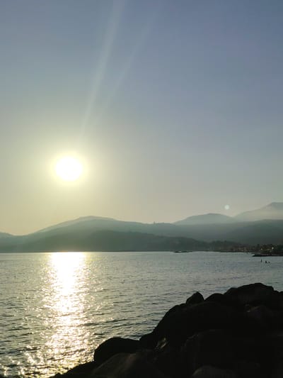 This vertical, outdoor photograph depicts a body of water with a shoreline and hills in the background, illuminated by the sun during what appears to be either sunrise or sunset. The sun, a bright, circular orb, is positioned in the upper left quadrant of the image, casting a strong reflection across the water's surface. The water is calm, with a subtle ripple effect, and the reflection of the sun creates a shimmering path of light extending towards the foreground.
In the mid-ground, silhouetted hills with a hazy appearance form a curved horizon. A town or village is faintly visible along the base of the hills on the right side of the frame, with small structures and lights suggested. The sky transitions from a pale yellow near the sun to a light blue towards the top of the image. Two faint, white lines extend diagonally from the sun upwards into the sky, possibly lens flares or atmospheric phenomena.
In the foreground, dark, irregular-shaped rocks occupy the lower right portion of the image, creating a stark contrast with the illuminated water. A few very small, dark figures are discernible in the water on the far right, too indistinct to be identified as people or objects, though they suggest the presence of activity in the distance. The overall scene conveys a sense of tranquility and natural beauty. The location is identified as Marina di Casal Velino, Italy.