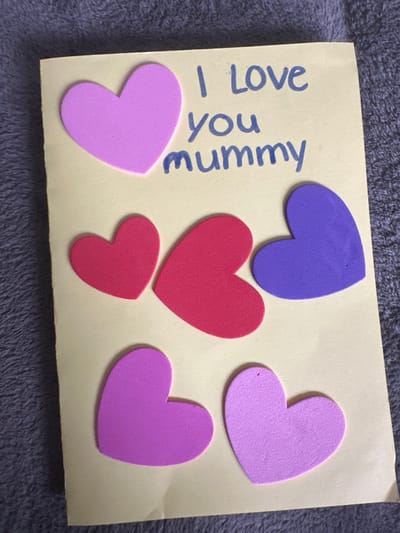 A handmade greeting card rests on a grey, textured fabric surface. The card is light yellow and features the handwritten phrase "I Love you mummy" in blue script at the upper right. Seven heart-shaped foam cutouts are adhered to the card's surface. These hearts are in varying shades of purple, pink, and red: one large light purple heart is positioned in the upper left, two red hearts and one dark purple heart are in the central area, and three additional light purple/pink hearts are arranged across the lower section. The card exhibits minor creasing, notably near the bottom right corner. This scene is located in Easingwold, United Kingdom.