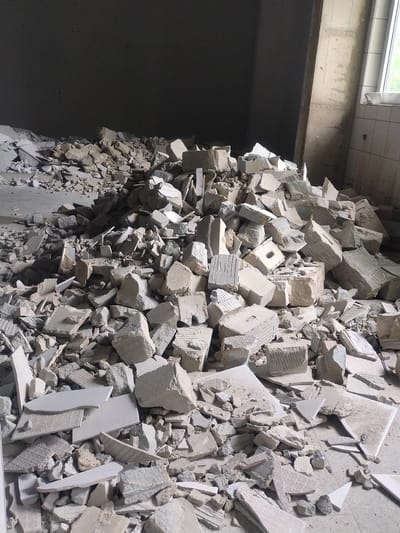 A large pile of construction debris, primarily broken concrete and light-colored tiles, dominates the right and central portions of the image. This scene, located in Bertrange, Luxembourg, strongly suggests a recent demolition or significant renovation project.

The debris is a chaotic mix of countless irregular fragments. Larger pieces are rough-hewn concrete blocks, some displaying textured surfaces. Interspersed are numerous broken ceramic or porcelain tiles, mainly in shades of off-white, light gray, and beige. Many tiles clearly show the ridged backing designed for adhesive, indicating their origin from walls or floors. Fine dust and grit are visible among the larger pieces, reinforcing the impression of recent activity.

In the upper-left, a robust concrete beam spans horizontally. Above it, a window or opening allows natural daylight to enter, illuminating the upper section of the debris and hinting at an outdoor environment beyond. To the right of the window, a light-colored surface with a grid pattern, possibly ceiling tiles, is visible.

A prominent, deep, and dark void occupies the entire left side of the image, providing a stark contrast to the brightly lit, textured debris. This dark area could be a lower floor, an empty room, or an unfinished space. The lighting indicates it is daytime. No people or visible text are present in the image. The overall impression is one of deconstruction and raw building materials.