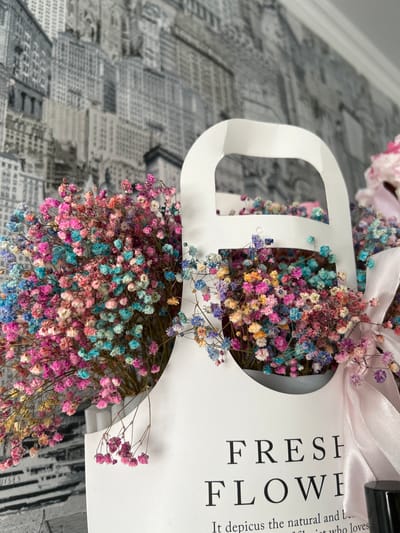A close-up shot presents a bouquet of colorful gypsophila flowers arranged in a white, handled container labeled "FRESH FLOWERS." The flowers exhibit a mix of pink, blue, yellow, and purple hues. A light pink ribbon is tied around the top of the container. The background features a black and white cityscape pattern. The location is unknown.