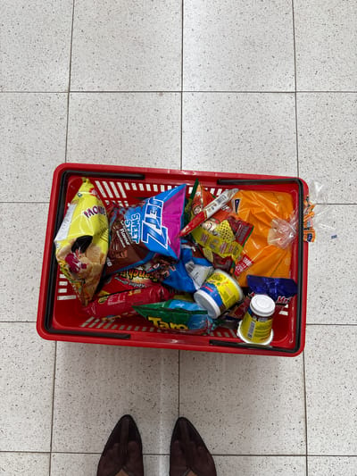 A red shopping basket is on a light gray tile floor, filled with various food items. These include a loaf of bread in an orange plastic bag, multiple cans and packages of snacks, including "JETZ Sweet Sticks Chocolate Flavour," "BONOVI," "Taro" chips, and "Kentang Goreng" chips. The basket is positioned near the edge of the frame, with a person's feet, shod in dark brown flats, partially visible to the right. The scene appears to be within a store in Sukabakti, Indonesia based on the product packaging language.