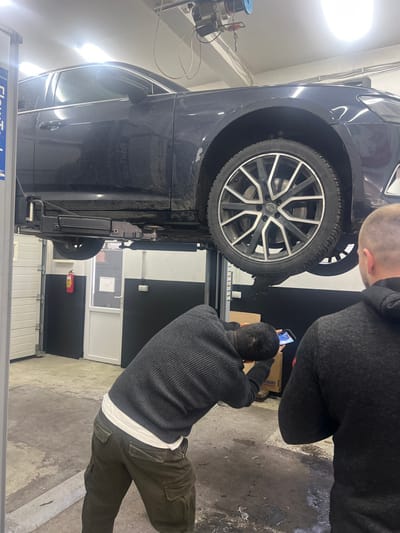 The scene depicts an interior view of an automotive service bay, likely located in Stara Pazova Municipality, Serbia. A dark-colored Audi sedan, appearing dirty on its lower half, is elevated on a hydraulic two-post vehicle lift. The vehicle's front passenger side wheel and fender area are prominent. Dark fluid appears to be dripping onto the concrete floor from the area beneath the front passenger side wheel.
Two adult males are present in the foreground. The man on the left, with his back partially to the camera and his head turned away, is bending over while holding a smartphone in both hands, actively photographing the underside of the elevated vehicle. He is wearing a dark ribbed sweater over a white t-shirt and dark olive green cargo pants. The man on the right, seen from the back of his head and shoulders, is observing the scene. He has short hair and is wearing a dark gray or black hooded sweatshirt.
The environment includes a concrete floor with visible stains and moisture. The walls are white with black panels along the lower section. To the left, a white roll-up garage door is partially visible. Further back and to the left, a white pedestrian door with an upper glass panel leads to another area. A red fire extinguisher is mounted on the wall near this door. Overhead, fluorescent lights illuminate the space, and a ceiling-mounted winch or hoist mechanism is visible.