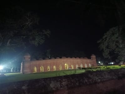 The image shows a historic brick building with arched openings, illuminated at night. The building appears to be a mosque or a similar structure, possibly with a dome. It is situated in a grassy area, with trees visible in the foreground and background. The sky is dark, indicating nighttime. The building's architecture suggests it is of historical significance. Based on common knowledge of the region, this building is likely the Bibi Begni Mosque in Bagerhat, Bangladesh, a UNESCO World Heritage Site known for its terracotta ornamentation and unique architectural style. The scene depicts a tranquil, atmospheric view of the illuminated structure under the night sky.