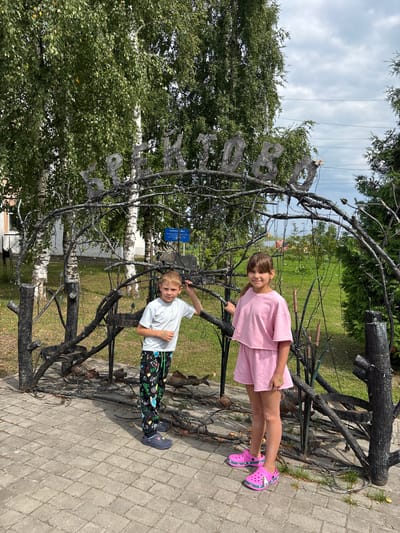The image depicts two children, a boy and a girl, standing near a decorative metal archway inscribed with the Cyrillic text "Брейтово 1050", indicating the town of Breytovo and its 1050th anniversary. The boy, wearing a white t-shirt and patterned pants, stands on the left, his hand resting on the arch. The girl, on the right, is dressed in a pink t-shirt and shorts. Behind them, birch trees and lush green grass form the background, with a cloudy sky overhead. The archway itself is constructed from dark, twisted metal branches, some adorned with small lights. A blue sign with Cyrillic writing is visible behind the arch, along with a metal sculpture of a bull or cow. The ground in front of the arch is paved with gray interlocking bricks. The overall scene suggests an outdoor public space, possibly a park or town square, celebrating a local anniversary.