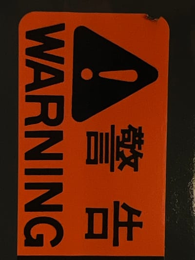 A vertical, orange sticker with black text and symbols is affixed to a dark, textured surface. The sticker has rounded corners and a small, chipped area on the top right edge. The text "WARNING" is printed vertically in large, black capital letters on the left side. To the right of "WARNING," a black equilateral triangle with a rounded tip and a dot inside is displayed. Below the triangle, two lines of black text in a different font are present. The top line consists of two Chinese characters, and the bottom line consists of two more Chinese characters. The scene is photographed in close-up, with the background out of focus, suggesting an indoor setting rather than a specific outdoor location. There is no indication that this image is located in Lanžhot, Czechia.