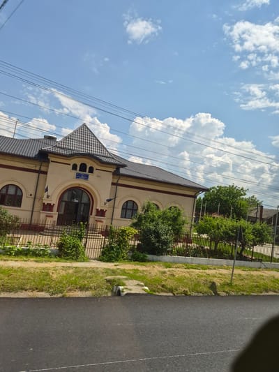 The image depicts the exterior of a light-colored, two-story building with a dark, tiled roof in Răcăciuni, Romania. The building features a prominent arched entrance with double doors and a small blue sign above it. Flanking the entrance are arched windows, and a Romanian flag is visible to the left of the entrance. A metal fence encloses the property, behind which are trees and lush green vegetation. Power lines crisscross the sky, which is a clear blue with scattered white clouds. The foreground shows a paved road and a grassy verge with a concrete drain. The scene appears to be captured on a sunny day.