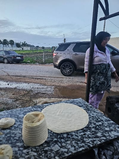 A woman in a headscarf and patterned clothing stands next to a vehicle, her hand resting on a granite surface. On the granite, there is a circular dough-like object, a rolled coil, and several smaller dough balls. The scene unfolds near a wet road with parked cars and a green field under a cloudy sky. An angled post frames the left side of the image.