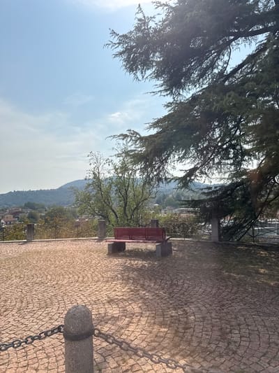 The image depicts an outdoor scene with a panoramic view of a town nestled in a valley, with hills in the background. In the foreground, a red bench is positioned on a cobblestone plaza, which is bordered by a low fence. A chain fence runs across the bottom left of the image, attached to a stone post. Large trees with dense foliage frame the upper right portion of the scene, casting shadows on the plaza. The sky is clear and blue with some light clouds. The location appears to be a viewpoint or park area, likely within or overlooking the town of Balerna, Switzerland, given the context. There are no people visible in the image.