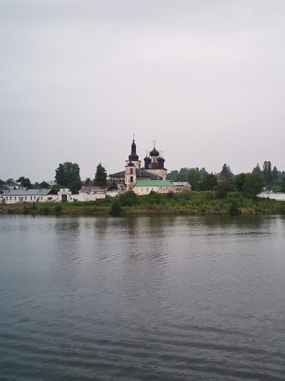 The image depicts the Goritsy Monastery, situated in Goritsy, Russia, on the shore of the White Lake. In the foreground, a wide, calm body of water, likely the White Lake itself, occupies the lower half of the frame. The water's surface is a muted, dark gray, with subtle ripples indicating a gentle current.
Dominating the background is the Goritsy Monastery complex. The architecture features multiple onion domes, some topped with crosses, characteristic of Russian Orthodox churches. The main church building is a prominent, multi-story structure with a mixture of aged stone and wooden elements. Adjacent to it are several other buildings, including white-washed structures with green roofs and various administrative or residential buildings. The monastery is nestled on a green, tree-lined embankment. Lush trees and dense foliage surround the monastery buildings, creating a verdant backdrop against the overcast sky. The sky itself is a uniform, pale gray, suggesting a cloudy or foggy day. There are no visible people or animals in the image. The scene is still and peaceful, capturing a historical and religious site.