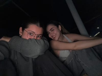 The image displays two young adult females seated in a dimly lit environment, likely at night. The specific location within Xcanatún, Mexico, cannot be determined from the visual evidence.
The individual on the left has dark hair pulled back and wears round-framed eyeglasses. She is dressed in a dark grey long-sleeved sweater and dark pants. Her left arm is bent, with the forearm and sleeve positioned horizontally across the lower half of her face, partially obscuring her nose and mouth. Her eyes are directed towards the camera. A thin, dark chain, possibly from a shoulder bag, is visible on her left side.
The individual on the right has lighter-colored hair, also pulled back, and is smiling with her teeth visible. She wears a light-colored halter-neck top and dark pants. Her body is angled slightly away from the camera, with her head turned to face it. Her left arm is extended forward and rests in front of the individual on the left.
The background is predominantly dark, suggesting a low-light setting. A light-colored, vertical support structure or pole is visible on the right side of the frame, extending from the bottom towards the top right. Some faint, blurred lights are discernible in the distant background. Both individuals are looking directly at the camera.
