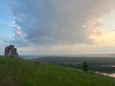 The image depicts a panoramic view from a grassy hill overlooking a valley. In the foreground, a partially ruined brick structure with an arched opening stands on the left side of the hill. The hill is covered in tall green grass with scattered white wildflowers. In the middle ground, the valley is densely populated with dark green trees, and a river or large body of water is visible on the right, reflecting the sky. The background consists of a hazy, undulating landscape under a dramatic sky filled with clouds. The sky transitions from a pale blue on the left to warm, soft hues of pink and yellow on the right, suggesting either sunrise or sunset. The scene appears to be set in an outdoor, natural environment, likely in or near the city of Nizhniye Vyazovye, Russia, given the characteristic landscape and vegetation. No people or animals are visible in the image. The overall impression is one of serene, expansive natural beauty with a touch of historical ruin.