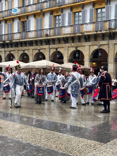 The image shows a group of people dressed in traditional military uniforms, playing drums, in a square in front of a building in Donostia, Spain, located at the coordinates (latitude: 43.324, longitude: -1.985). The building has a yellow facade and is decorated with white window frames and black balconies. The square is paved with grey stone tiles, and the drums have a red and blue design with the word "AMBROSIO" written on them. The people are in a parade formation, and are all wearing hats, and some are also wearing white ponchos. The square is wet, suggesting that it has been raining. The sky is not visible in the picture.