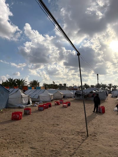 An outdoor scene in a-Zawayda, Palestinian Territory, depicts a temporary settlement under a partly cloudy sky with prominent sunlight from the upper right. The foreground and mid-ground consist of light brown sand, showing numerous footprints. A dark-colored, possibly metal, utility pole stands diagonally in the foreground, extending upwards and to the right, with dark wires strung across the upper portion of the frame. A second, similar pole is visible further back, supporting the wires.

The central and background areas are occupied by multiple rows of white or light-colored canvas tents, varying in size and apparent construction, indicative of a displacement camp. Palm trees are densely clustered behind the tents, especially along the horizon line on both the left and right sides of the image. Between the rows of tents, a pathway of sand is visible.

In the mid-ground, approximately ten red, rectangular objects, resembling large bags or containers, are lined up on the sand, extending from the left towards the center. On the right side of the mid-ground, a single individual, dressed in dark clothing (dark jacket, dark pants), stands facing away from the camera, next to another similar red object. Clothes are visibly hanging to dry on a line outside a tent further back on the right. White sacks or bundles are visible on the ground near the entrance of a tent on the far left. Shadows from the poles, tents, and the individual are cast towards the left and foreground, consistent with the sun's position.