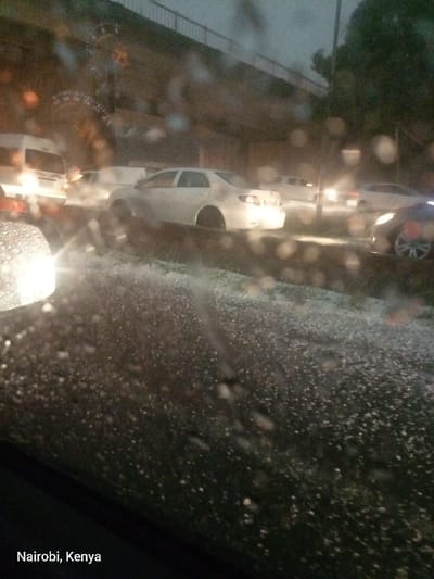 The image depicts a nighttime or dusk scene on a street in Benoni, South Africa, during or immediately after a hailstorm. The perspective is from inside a vehicle, looking out through a windshield covered in raindrops and small hail, which blurs the scene and creates circular bokeh effects from light sources.
The ground in the foreground is heavily covered with small, white hail pellets, clearly indicating severe weather conditions. Several vehicles are visible, suggesting traffic. A prominent white sedan with illuminated taillights is in the midground, its tires almost obscured by the hail accumulation. Behind it, a white van or SUV is partially visible, along with other cars whose headlights and taillights pierce through the dim, wet environment. To the far right, a dark car is partially seen. An elevated structure, likely an overpass or bridge, stretches across the upper part of the frame, hinting at a multi-level urban infrastructure. The artificial lights from vehicles and possibly streetlights are the primary sources of illumination against the dark, overcast sky. The overall impression is one of difficult driving conditions due to the heavy hail and rain, compounded by low visibility. The text "Nairobi, Kenya" is visible in the bottom left corner of the image.