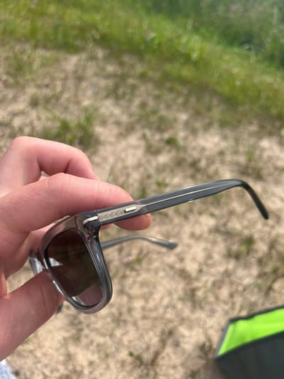 A person's hand is holding a pair of translucent grey Gucci sunglasses outdoors. The sunglasses have dark lenses and a small silver Gucci logo on the temple. The background is blurred and consists of sand and grass, suggesting an outdoor setting in Vangaži, Latvia. The lighting indicates it is daytime.