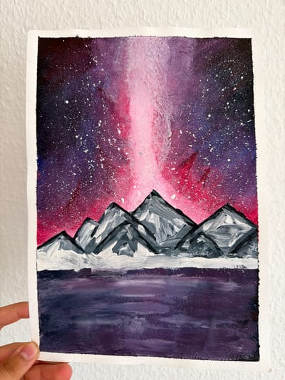 The image shows a watercolor painting of a mountain landscape under a vibrant night sky. The sky is a blend of deep purples and bright pinks, with white specks representing stars. A prominent, light pink aurora-like beam stretches vertically through the sky. In the foreground, silhouetted mountains with snow-capped peaks are depicted in shades of gray and white. Below the mountains is a dark, shimmering body of water, painted in shades of deep purple and black. The painting is on a textured white paper with a torn edge on the right side. A hand is holding the bottom left corner of the painting, revealing fingernails and a portion of the thumb. The background of the image is a textured white wall. There is no discernible location in Nienburg/Weser, Germany depicted in this artwork.