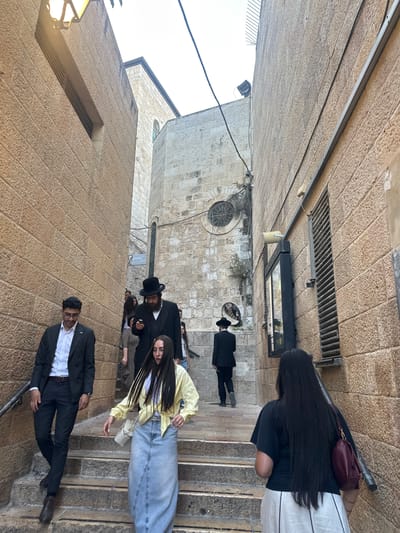 The image depicts a group of people in an outdoor, narrow, stone-walled passageway. The architecture suggests an ancient or historic urban setting. The people are dressed in a mix of modern and traditional attire. Some individuals wear black suits and hats, possibly indicating a religious or formal occasion, while others are in casual modern clothing like a yellow jacket and jeans.
The scene appears to be a street or alleyway with stone paving and walls. There are stairs visible in the foreground and background, suggesting a hilly or terraced terrain. A street lamp is visible on the left side of the image, and a circular window with a decorative grating is set into a stone building on the right.
The people are walking, some looking at their phones, others interacting or looking around. The mood seems to be one of everyday activity, with no overt signs of distress or celebration. The lighting suggests it is daytime, possibly late morning or afternoon, with no strong shadows indicating harsh sunlight. The overall atmosphere is calm and ordinary.
No visible text is discernible in the image. The location context of al-Eizariya, Palestinian Territories, points to a Middle Eastern setting, which aligns with the stone architecture and the traditional attire of some individuals.