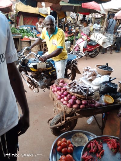 A bustling market scene unfolds with a man on a motorcycle in the foreground. A cart laden with onions, dried fish, and bagged goods stands nearby. Metal plates hold tomatoes and chili peppers in the immediate foreground. Behind the man, a glimpse of vendors and shoppers amongst stalls, some shaded by umbrellas, reveals a vibrant trade environment. Another red motorcycle is parked further back.