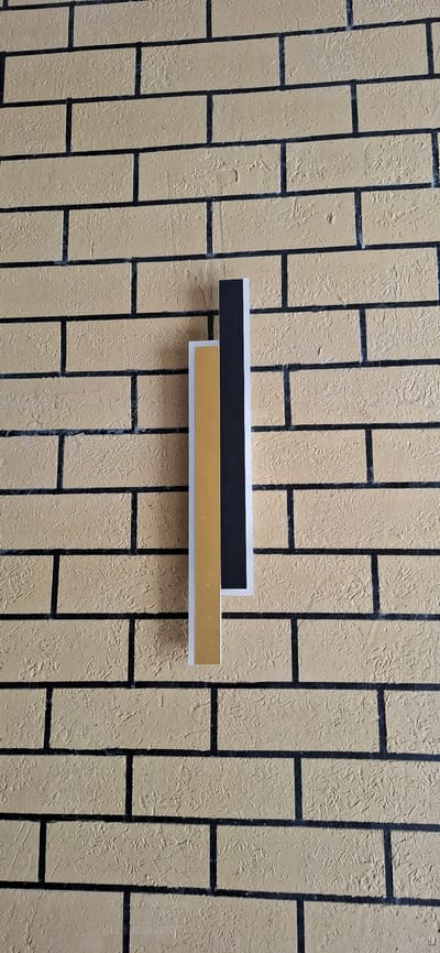 The image shows a close-up, head-on view of a textured, tan-colored brick wall with black mortar lines. Mounted vertically on the wall are three rectangular decorative elements: one is a thin, silver-colored strip on the left; next to it is a wider, golden-colored strip; and to the right of that is a thin, black strip. The elements appear to be mounted close together, aligned vertically. The scene is indoors, likely in a public space or a home in the city of Karmana, Uzbekistan. There are no people or animals present in the image. The lighting is even, suggesting artificial light.