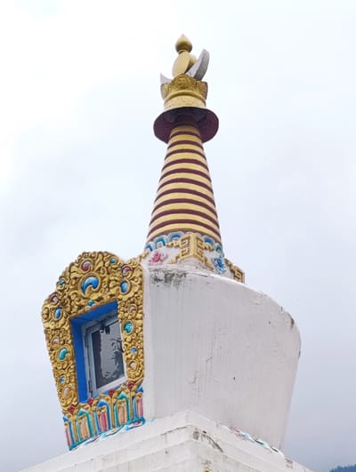 The image shows a close-up, low-angle shot of the top section of a stupa, a Buddhist monument, against an overcast sky. The stupa is predominantly white with colorful decorative elements. The uppermost part is golden and conical, with horizontal red and gold stripes, topped by a golden finial. A distinctive feature is a recessed window framed by an ornate, golden, curvilinear structure adorned with colorful gemstones in shades of blue, red, and teal. Below the window, there are tiered, decorative bands in vibrant colors, including blue, red, and yellow. The white surface of the stupa shows signs of wear and peeling paint. The lower part of the stupa is stepped and also displays some peeling paint and faded colorful details. The background is a uniformly grey sky. The image does not contain any people or visible actions. The location is identified as Gadherni, India, which is a known area for Buddhist stupas and monasteries.