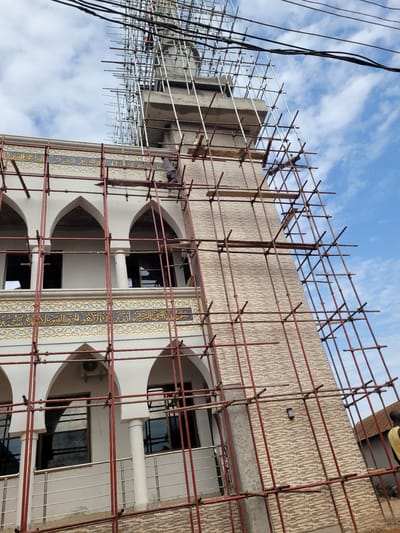 The image shows a multi-story building in Nassarawa Egon, Nigeria, under construction or renovation, as indicated by the scaffolding surrounding the facade. The building features arched windows and decorative elements, including panels with Arabic script. Construction workers are visible on the scaffolding. The scaffolding itself is made of metal poles and crossbars. Wires are visible in the upper left corner. The sky with clouds is visible in the background. The scaffolding covers almost all of the walls except for one corner on the far right.