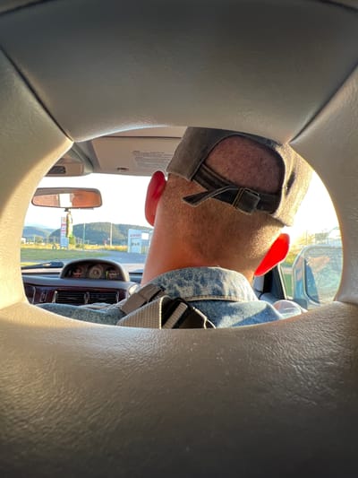 The image is taken from the backseat of a car, looking forward through a circular opening formed by the car's interior. The focus is on the back of the head and shoulders of a man wearing a denim jacket and a baseball cap. His ears are red, likely from the sun. The dashboard of the car is visible, including gauges and vents. Outside the car, through the windshield and side windows, a rural landscape with buildings, hills, and trees can be seen. A gas station with a sign that reads "SHELL" is visible in the distance, suggesting the car is near a road. The lighting indicates it is late afternoon or early evening, with a warm, golden light illuminating the scene. The location is identified as Dragushinovo, Bulgaria.