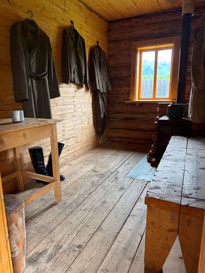 This is an interior shot of a rustic wooden room, likely a cabin or barrack, located in Ust'-Kabyrza, Russia. The room is sparsely furnished, emphasizing practicality and functionality.

On the left wall, three military-style jackets are hung on hangers. The leftmost jacket is a long overcoat, cinched with a belt, and has red accents on the collar. To its right, a shorter jacket is also hanging, followed by a third, similar garment. Below these, a simple wooden table stands, supporting a metal mug. Beneath the table, a pair of tall black boots are placed on the wooden floor.

The right side of the room features a wood-burning stove with a dark metal chimney pipe extending upwards. A green metal kettle sits atop the stove. In front of the stove, a stone or metal mat is laid on the floor. A rough-hewn wooden bench occupies the foreground on the right.

A single wooden-framed window is visible on the back wall, offering a view of a green, hilly landscape with a wooden fence. The walls and floor are constructed from rough-hewn wooden planks, giving the space a primitive and utilitarian appearance. The lighting is natural, coming from the window and an unseen source, creating shadows that highlight the textures of the wood. The overall impression is one of a functional, living space, possibly from a historical military or frontier context.