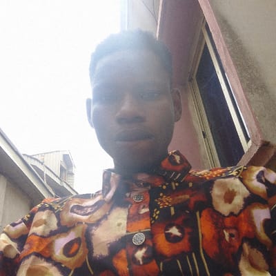 A young, dark-skinned man is captured in a low-angle selfie. He wears a brown and orange patterned shirt with two visible silver buttons. The background shows a mix of building exteriors, including what appears to be a window with dark frames and parts of a wall with varying shades of color. The sky is bright and overcast. The overall lighting creates shadows on the man's face and emphasizes the textures of his clothing.