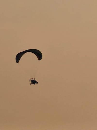 A paramotor is captured mid-flight against a muted, hazy sky. The paramotor, appearing as a dark silhouette, consists of a pilot harness with visible struts and a propeller, attached to a dark, curved wing that billows above. The wing's dark outline is sharply defined against the lighter, uniform background. The paramotor is positioned in the lower-middle portion of the frame, with the wing arching overhead and to the left. The overall impression is one of solitary flight in an atmospheric, possibly dusty or overcast, environment. The specific location in Lesosibirsk, Russia, cannot be determined from the visual information provided in the image.