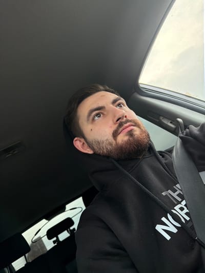A male individual is depicted from the chest up, seated in what appears to be the driver's seat of a vehicle. He has short dark hair, a dark beard, and a mustache. His brown eyes are directed upwards and to the viewer's right. He wears a black hooded sweatshirt featuring the white "THE NORTH FACE" logo on the chest. A black seatbelt is positioned diagonally across his left shoulder and chest. The vehicle's interior includes a dark gray headliner and a partially open sunroof on the right side of the frame, revealing an overcast sky. A rearview mirror is partially visible in the lower left background. The specific location within the city of Zlatina, Bulgaria, cannot be determined from the visual evidence.