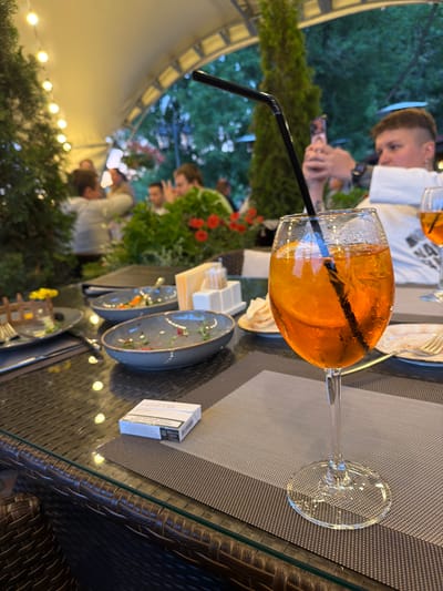 The image depicts an outdoor seating area of a restaurant or cafe, likely in Izmailovo, Russia, as indicated by the specific context of the prompt. The scene is set at a table with a glass-top surface, adorned with woven placemats. In the foreground, a tall stemmed glass filled with an orange-colored beverage, garnished with a slice of orange and a black straw, is prominently featured. Beside it, a pack of HEETS cigarettes rests on a placemat.
The table is also set with several used dishes, suggesting a meal has been or is being consumed. One plate contains remnants of food and scattered greenery. Another deeper bowl holds a small amount of salad or vegetables. A cutlery set and a small plate with what appears to be bread or crackers are also visible.
In the background, blurred figures of people are seen seated at tables under a white tent-like canopy, illuminated by a string of lights. Trees and foliage frame the outdoor setting. One person in the mid-ground is holding up a mobile phone, possibly taking a photograph. The overall atmosphere suggests a relaxed, social gathering during daylight hours, possibly late afternoon or early evening, given the lighting.