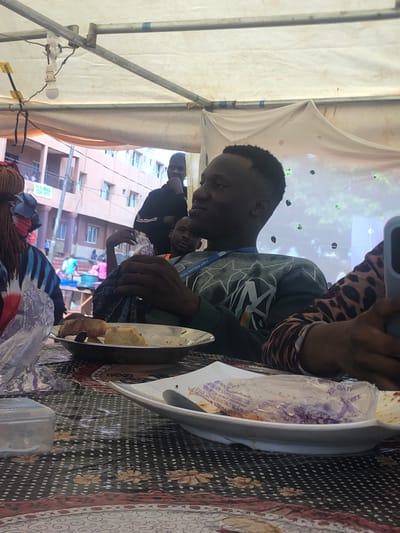 The image shows a group of people eating a meal under a tent-like structure. A man in a green sweatshirt with an orange lanyard around his neck is the main focus, seemingly engaged in eating. He is seated at a table covered with a patterned tablecloth. Another person with a leopard print sleeve is partly visible next to him, holding what appears to be a phone or tablet. In the background, other individuals are visible, along with buildings, hinting at an urban environment. A building with a sign is visible with words: ST PAULS TO, WEJE UMUNYA ROAD NIEJE, ANAMERA SATGERIA. From the text, it is determined the location is near Weje Umunya Road in Nteje, Nigeria.