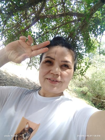 A woman with dark hair, styled in a bun, is taking a selfie outdoors. She is wearing a white t-shirt with a graphic of a cat's face on the front. Her right hand is raised, holding up two fingers in a peace sign. She is looking towards the camera with a slight smile. The background shows lush green foliage from a tree overhead and a sunlit, dry, unpaved area. The timestamp on the image indicates it was taken on June 21, 2026, at 13:25. The overall impression is a casual, outdoor portrait. The location is identified as Aktash, Uzbekistan.