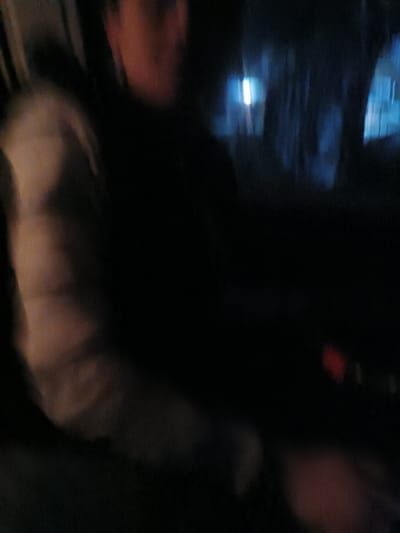 A blurry and dark indoor shot features a person's upper body. They wear a light-colored, possibly quilted, jacket. The face is partially obscured but seems to be in shadow. Outside, there's a glimpse of a dimly lit street scene through a window or windshield, showing trees and a blurred light source. The overall impression is of a nighttime or poorly lit environment within a vehicle or room.