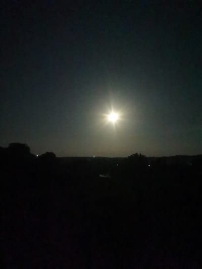 A nighttime landscape scene features a bright, starburst-like moon in the upper center of the frame against a dark, gradient sky. The moon is the most illuminated object in the image. Silhouetted against the sky is a dark, undulating horizon line with the indistinct shapes of trees and possibly hills. A faint, dark body of water may be present in the lower portion of the scene. The overall impression is one of a rural or undeveloped area in the city of Cărbuna, Moldova, under a moonlit sky.