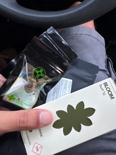 A hand holds a clear plastic bag containing cannabis. The bag has a green and black logo depicting a sun symbol with radiating lines, and text that reads "DISPENSARY NEAR ME". Another, darker bag is visible behind it. To the right, a white cardboard box with a cut-out flower design displays the text "BLOOM Surf 1g". The scene appears to be inside a vehicle, with a portion of a steering wheel visible at the top and gray fabric, likely clothing, in the background. The image was captured in Sunland Park, New Mexico, United States of America.