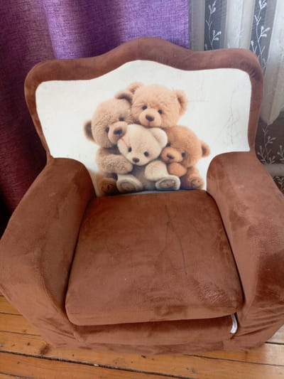 A brown, plush children's armchair with a printed image of three teddy bears is positioned on a wooden floor. The armchair has a high, curved backrest with a white panel featuring the teddy bear illustration. Behind the armchair, a purple curtain is visible on the left, and sheer white curtains with a floral pattern are on the right. The armchair appears to be in a domestic setting, possibly a living room or playroom. The image does not provide any information related to the city of Churapcha, Russia.