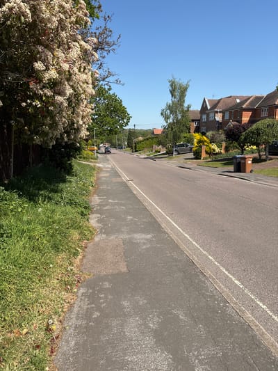 The image depicts a residential street in Welwyn Hatfield, United Kingdom on a sunny day. The road is asphalt, with a white painted line delineating the edge. A sidewalk runs alongside the road, bordered by grass and various plants. Houses are visible on the left side of the street, including brick structures with gabled roofs. One car is parked along the roadside. Trash bins are placed near one of the houses. A flowering bush is in the foreground. The sky is clear and blue.