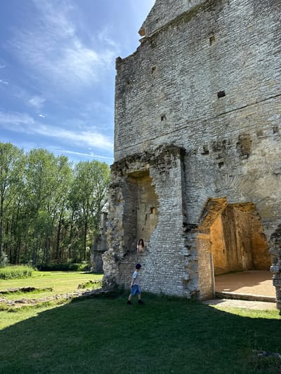 The image captures a scene at Minster Lovell Hall in West Oxfordshire, United Kingdom. A partial view of the ruin is seen. Two people are standing next to the ruined wall. Grass covers the ground. Trees are visible in the background. The sky is blue with scattered clouds.