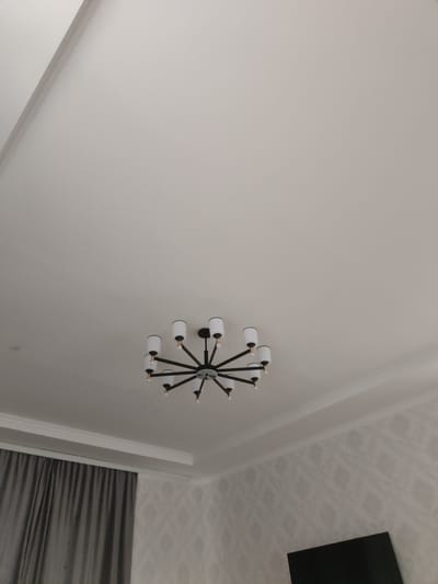 The image shows an interior view of a room with a white ceiling and textured walls. A black chandelier with eight white light fixtures is suspended from the center of the ceiling. In the lower left of the image, a portion of a gray curtain is visible. In the lower right, the top edge of a dark-colored electronic screen, likely a television, is present. The scene appears to be a domestic setting, possibly a living room or dining area. The image does not provide information about the specific location within Жарлыкөл, Kazakhstan, other than it is an indoor scene.