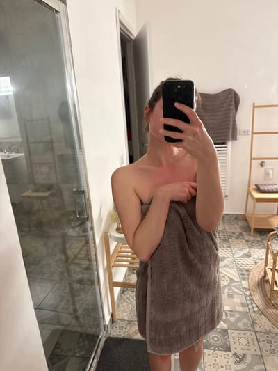 A person is taking a selfie in a bathroom. The person is holding a black smartphone, obscuring their face. They are wrapped in a gray towel. The bathroom features a glass shower door, patterned floor tiles, a wooden shelving unit, and a towel rack. The specific location in Perugia, Italy cannot be determined from the visual information alone.