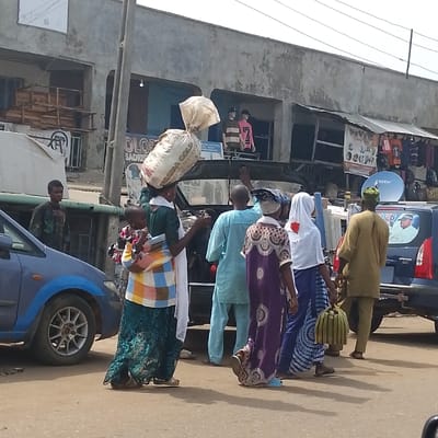 The image captures a bustling street scene in Olomoda, Nigeria, under bright daylight with visible shadows. In the foreground, a woman in a green top and a long patterned skirt walks from right to left, carrying two large white sacks on her head and a child strapped to her back with a colorful fabric carrier. Ahead of her, a group of five individuals, mostly with their backs to the viewer, are moving or standing. This group includes a man in a light blue tunic, a woman in a purple patterned dress, a woman in a white headscarf and blue patterned skirt carrying a green striped bag, and a man in a light brown tunic and green cap. On the far left, a man in a dark patterned shirt stands beside a partially visible blue sedan.
Vehicles populate the street: a blue sedan on the left, a dark-colored vehicle centrally positioned behind the pedestrians, and a blue car on the far right displaying a "2026" sticker with a political portrait and a satellite dish mounted on its rear. The background features two-story concrete buildings, some with visible wear, housing various shops. Merchandise, including clothing displayed on mannequins and hangers, is visible in the storefronts. Overhead electrical wires and poles traverse the scene.